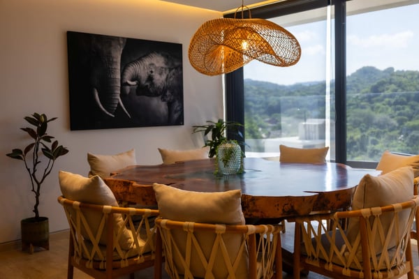 Modern dining room with round wood table, woven pendant light, mountain views through floor-to-ceiling windows, and elephant artwork on wall