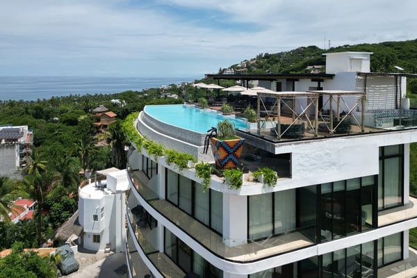 Modern dining room with round wood table, rattan chairs, woven pendant light, elephant artwork, and panoramic mountain valley views through floor-to-ceiling